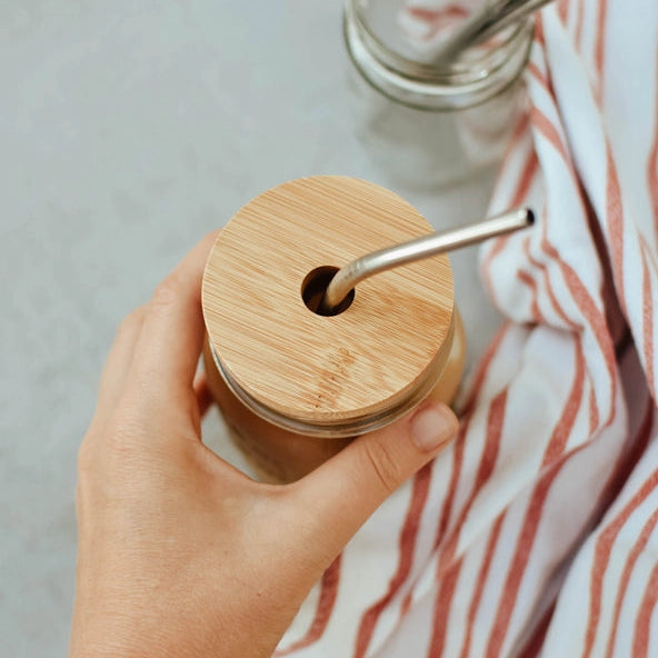 Hand holding a glass jar with a bamboo lid and metal straw, with a striped cloth in the background.